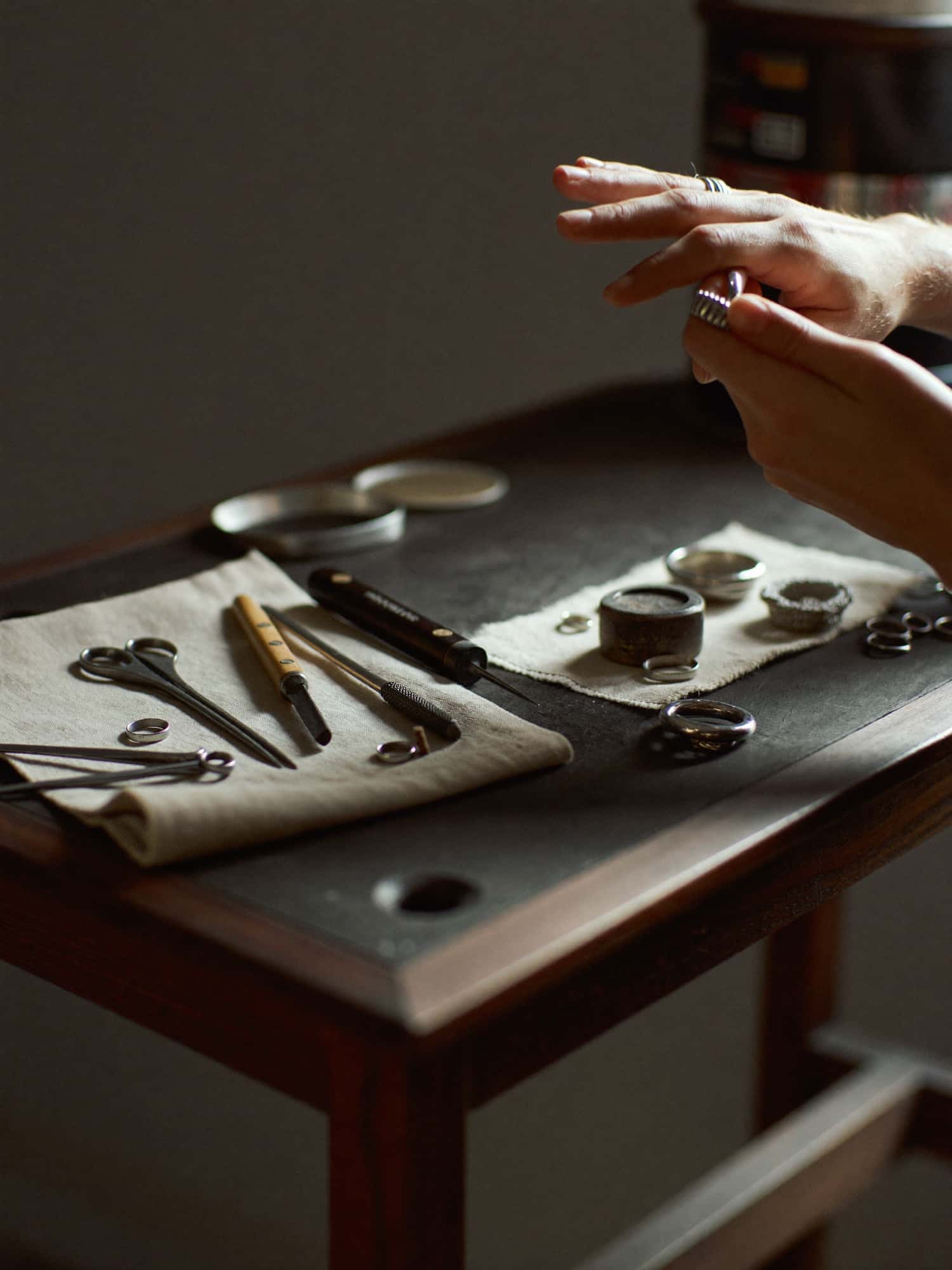 A silversmith's hands assembling silver rings among tools on a dark workbench